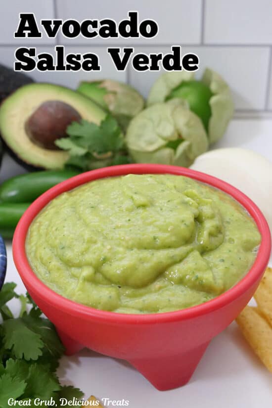A red bowl filled with creamy avocado salsa verde with the ingredients used placed in the background.