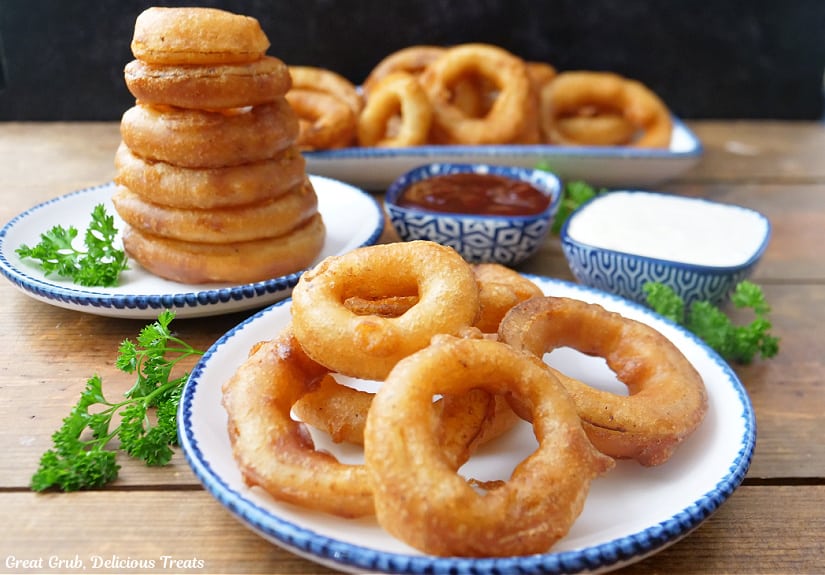 A horizonal photo of beer battered onion rings on three white plates with blue trim with two small blue bowls filled with BBQ sauce and ranch dressing.