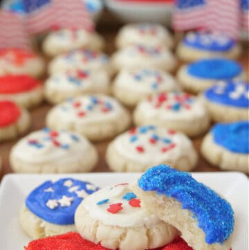 A white plate with mini red, white and blue sugar cookies on it with more cookies n the background.