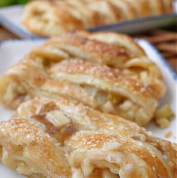 Three slices of apple puff pastry on a white plate with a whole puff pastry in the background on a white plate.