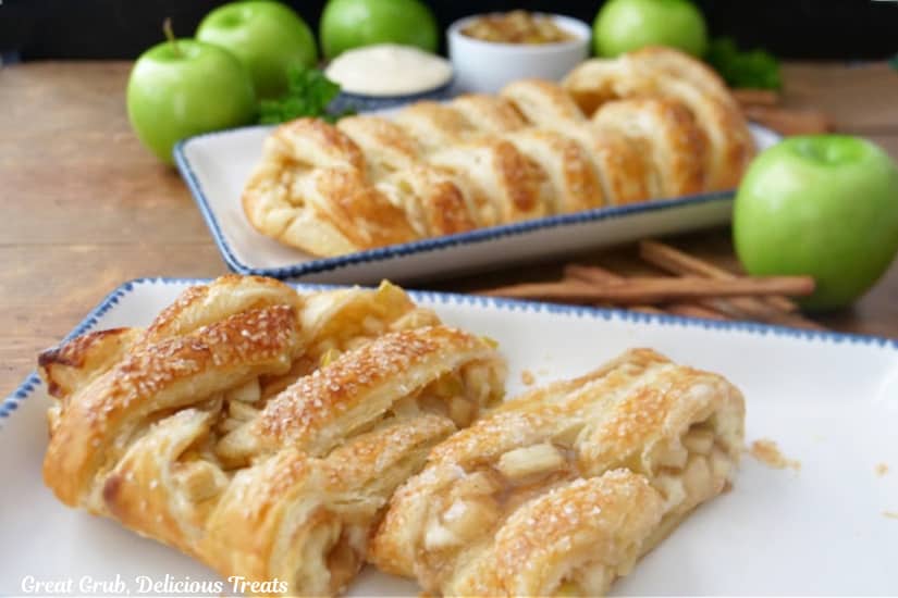 A horizontal photo of two apple puff pastry braids on white plates with blue trim with green apples in the background.