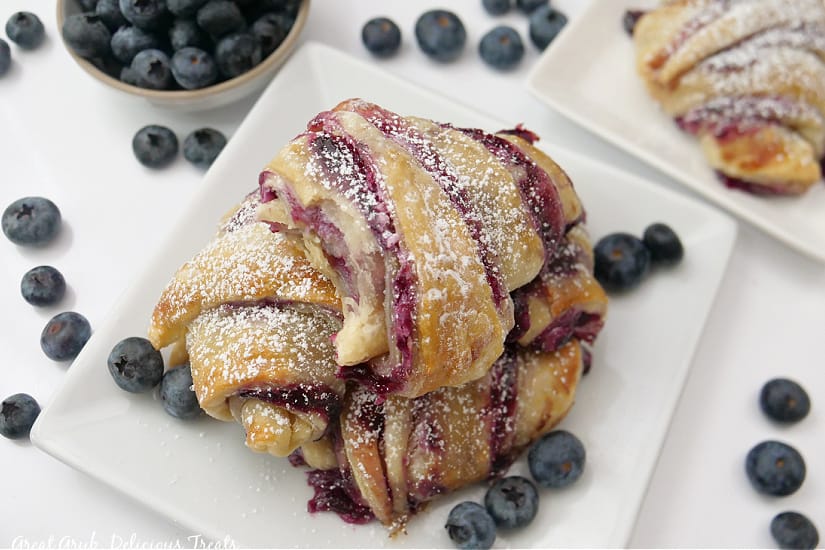 A horizontal photo of three blueberry pastries on a white plate with loose blueberries on the white surface and plate.