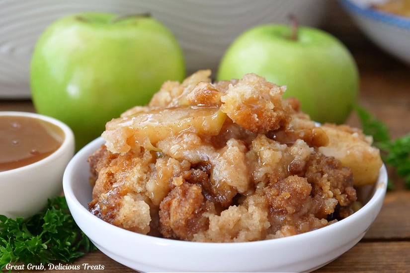 A horizontal photo of a white bowl filled with apple cobbler with two green apples in the background.