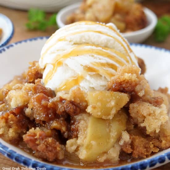 A white bowl with blue trim filled with caramel apple cobbler with vanilla ice cream on top.