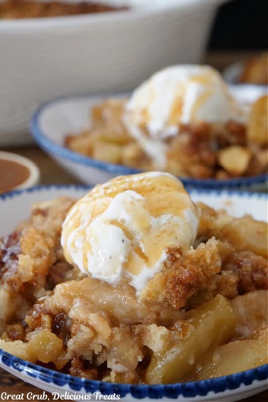 Two white bowls with caramel apple cobbler and a scoop of vanilla ice cream on top of each, drizzled with caramel.