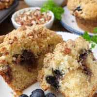 A white plate with a coffee cake muffin on it that was cut in half, and blueberries on the plate too.