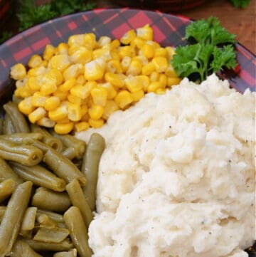 A red and black plaid plate with mashed potatoes, corn and green beans on it.