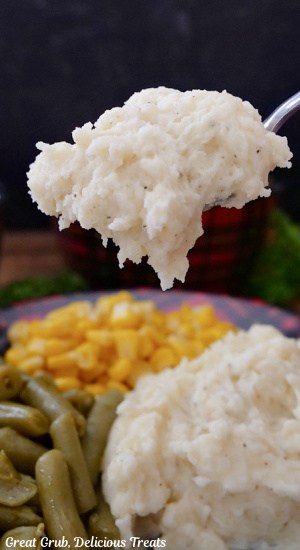 A big spoonful of mashed potatoes held above a plate with corn and green beans on it.