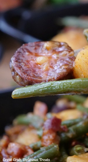 A close up of a piece of sausage, green beans and potatoes on a fork.