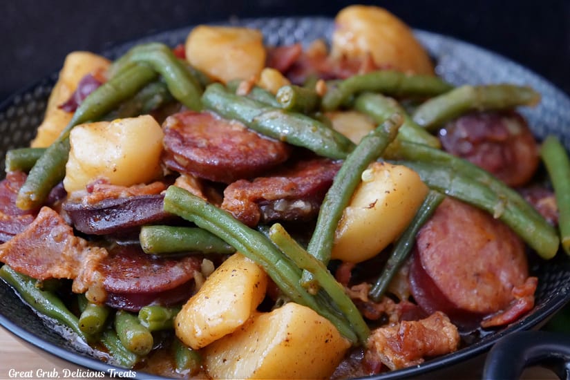 A horizontal photo of a black bowl filled with sausage, green beans, potatoes and bacon.