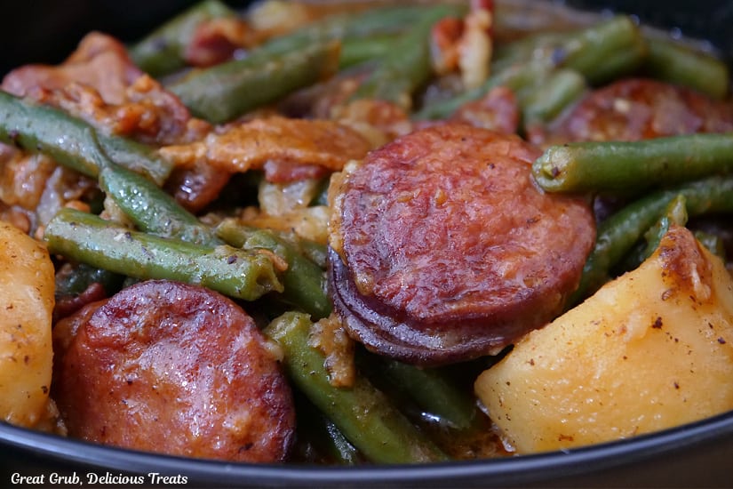 A close up of sausage, potatoes, green beans and bacon in a black bowl.
