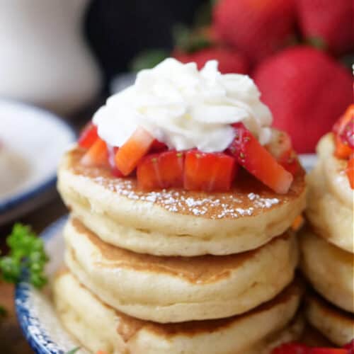 A white plate with blue trim with mini pancakes on it that are topped with strawberries and whipped cream.