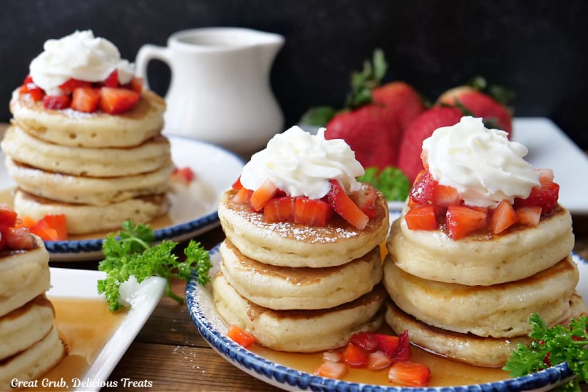 A horizontal photo of mini pancakes topped with strawberries and whipped cream.