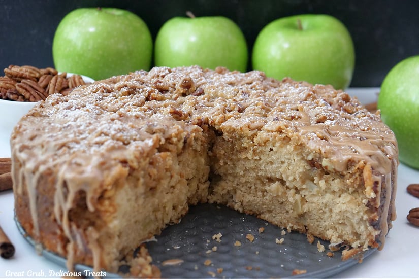 An apple coffee cake with a few slices removed, with green apples and pecans in the background.