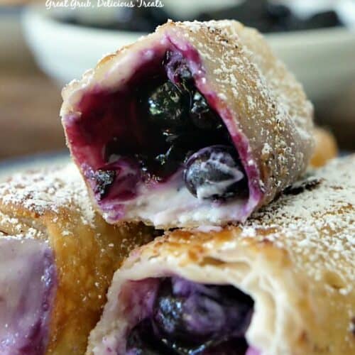 Three blueberry egg rolls on a white plate with blue trim showing the filling inside.