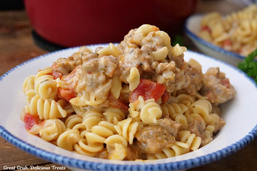 A horizontal photo of a white bowl with blue trim filled with a serving of sausage, pasta, tomatoes, and cheese.