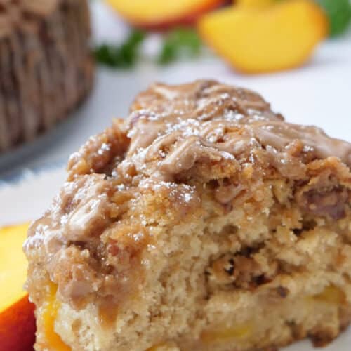 A slice of peach coffee cake on a white plate with peaches in the background.