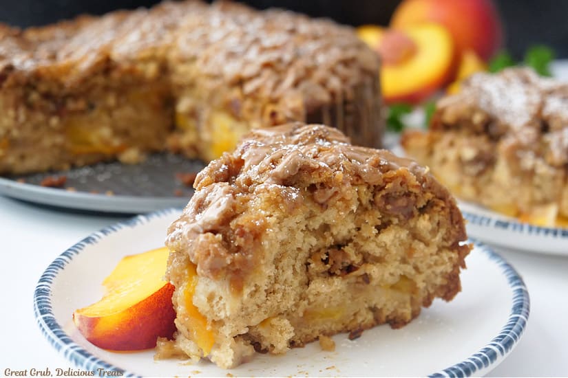 A horizontal photo of a slice of peach coffee cake on a white plate with blue trim and the coffee cake in the background.