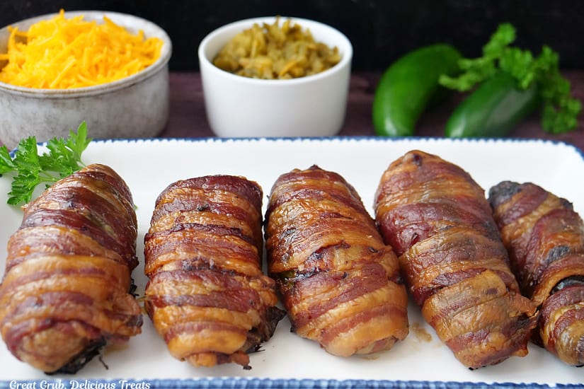A horizontal photo of a white oblong plate with blue trim with five bacon wrapped stuffed jalapeno poppers.