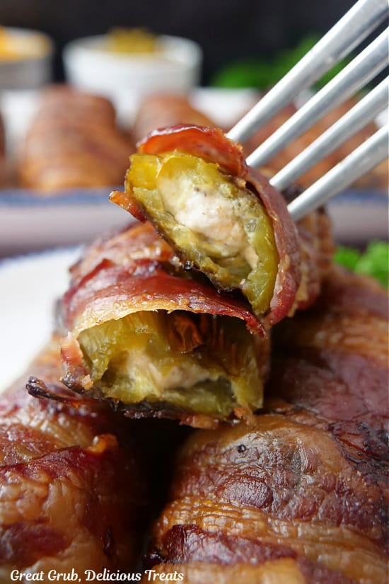 A white plate with three bacon wrapped jalapeno peppers with one cut in half and a fork pushed into it.