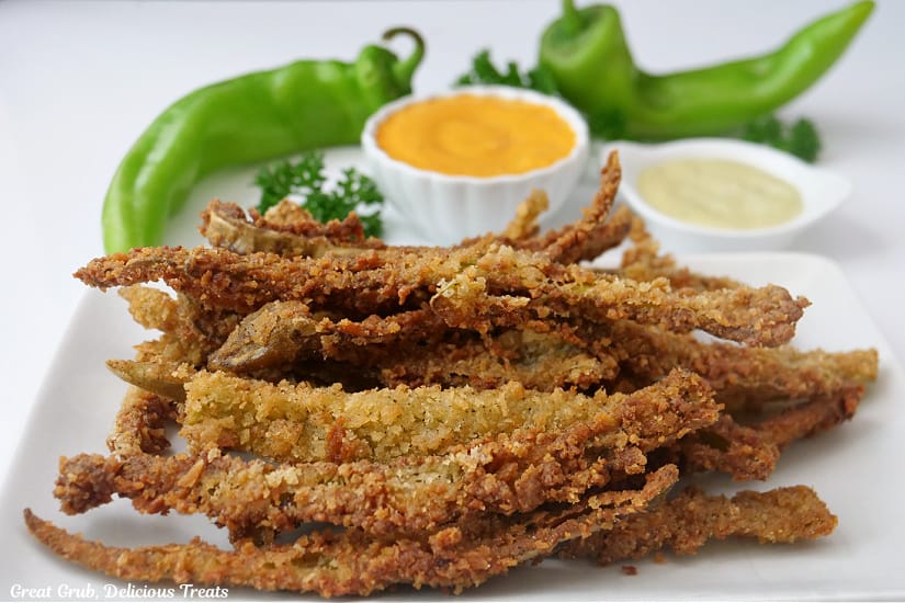 A horizontal photo of a white surface with a white plate with crispy deep fried hatch chile strips with two hatch chiles in the background with two small bowls with dipping sauce.