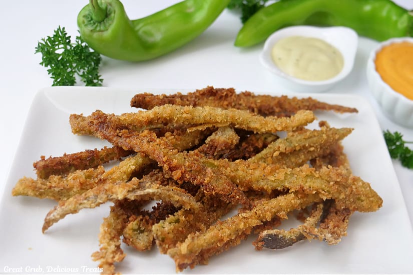 A horizontal photo of a white plate with deep fried hatch green chile fries on it with hatch chile peppers in the background and two small bowls with dipping sauce in them.