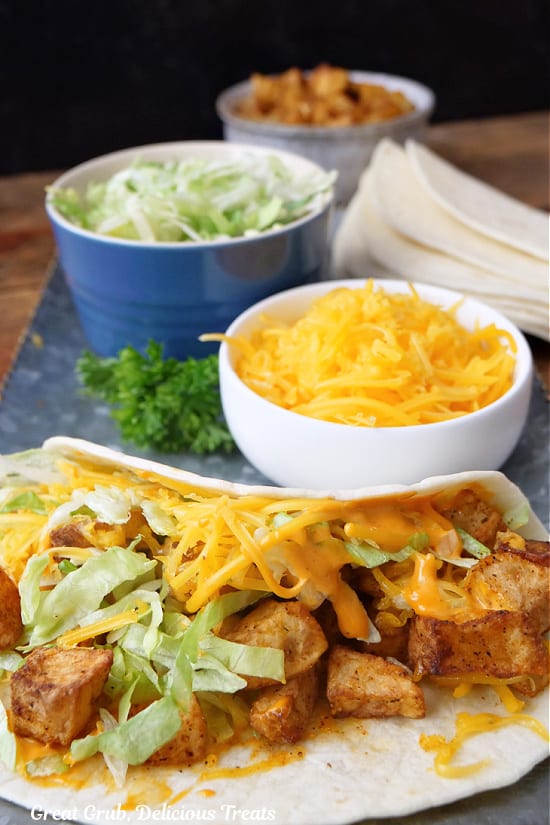 A sliver plate with a potato taco on it with a small bowl of cheese, lettuce and diced potatoes.