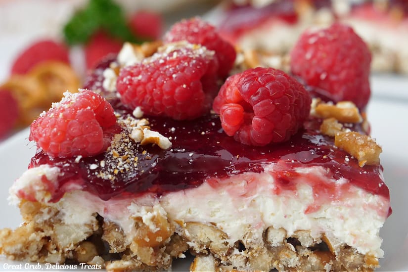 A horizontal photo of a close up of a slice of dessert with fresh raspberries, cream cheese, and pretzel crust.