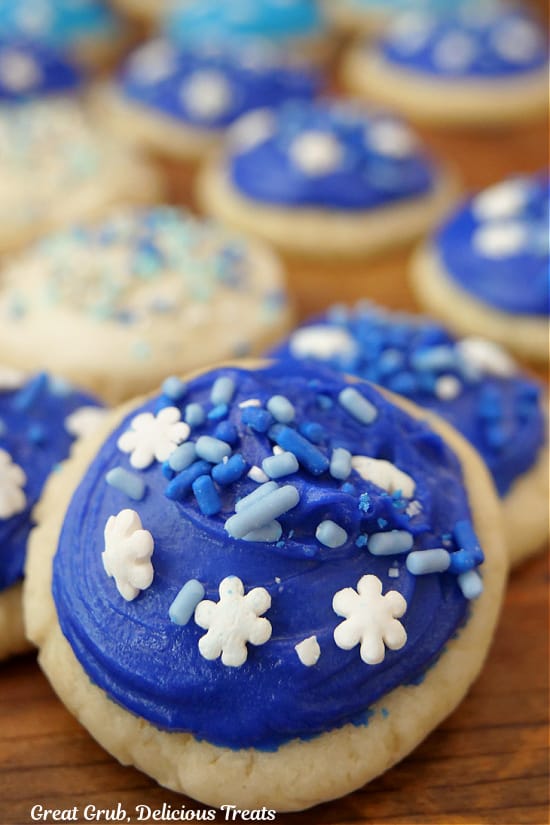 A close up of a mini sugar cookie with blue frosting and festive candy sprinkles on top with more cookies in the background.