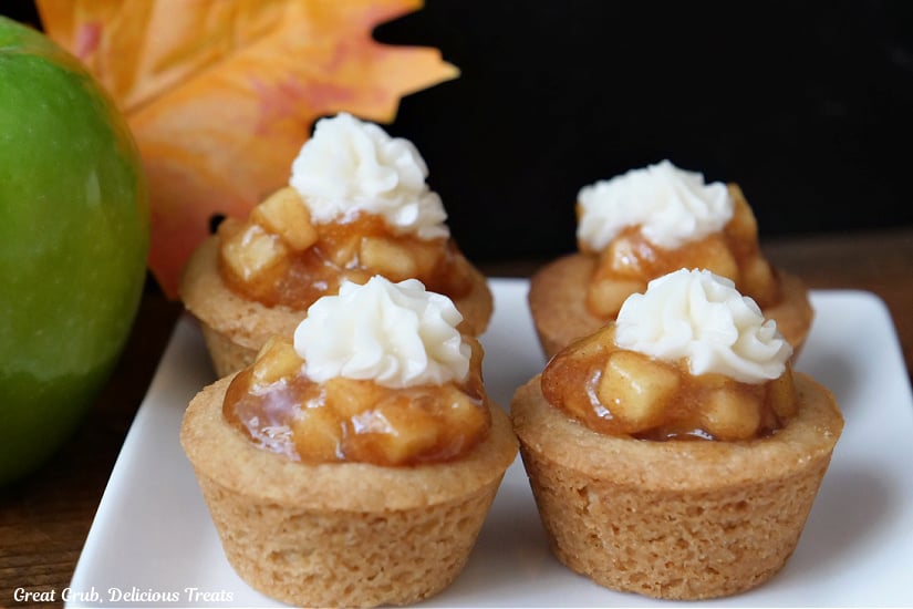 A horizontal photo of a white plate with four mini cookie cups on it that are filling with apple pie filling.