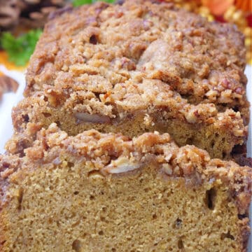 A loaf of butternut squash quick bread with a crunchy topping, sliced and has fall foliage in the background.