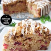 A white plate with a slice of cranberry coffee cake on it with the whole cake in the background.