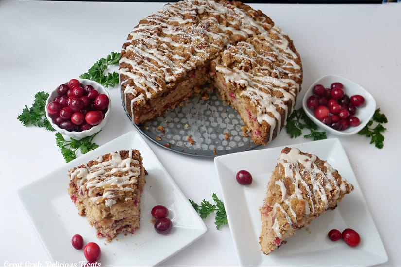 An overhead photo of two slices of coffee cake on two white plates with two small white bowls filled with fresh cranberries, and the rest of the coffee cake.