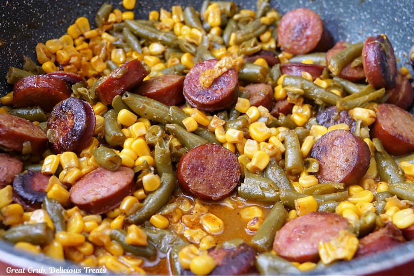 A horizontal photo of a skillet with sliced sausage, green beans and corn.