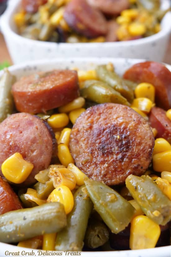 A close up of sliced smoked sausage, green beans and corn in a small white serving bowl.