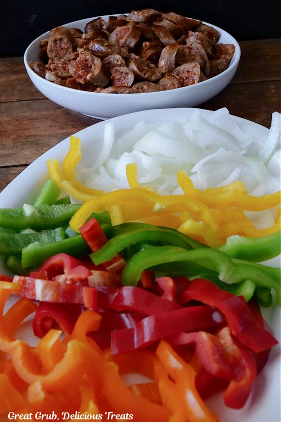Cooked sliced Italian sausage in a white bowl, and sliced onions, yellow, green, red, and orange bell peppers of a white plate.