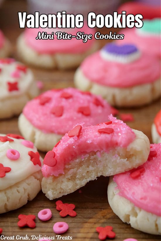 A few bite-size sugar cookies with pink and white buttercream frosting and candy sprinkles on top, and one with a bite taken out of it.