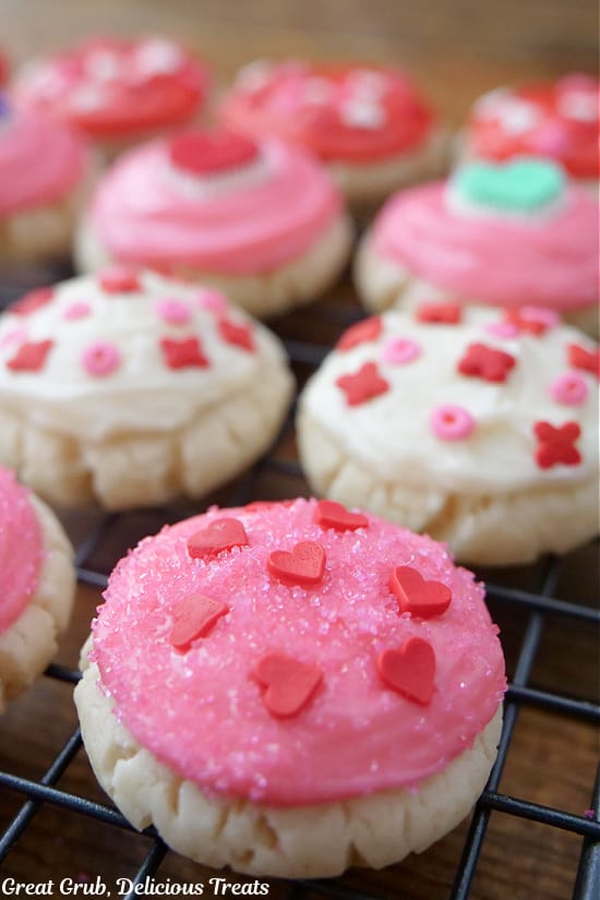 A close up of a few sugar cookies on a wire rack that are decorated with pink, white, and red frosting and candy sprinkles on top.