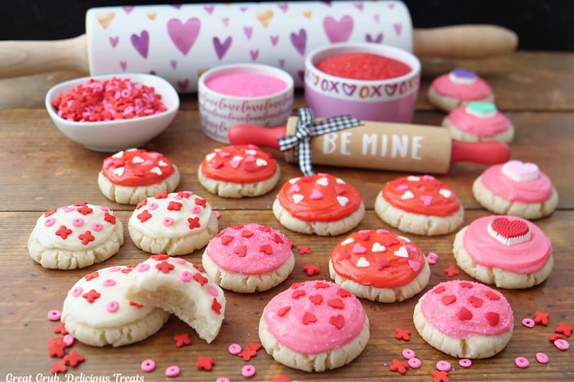 A horizontal photo of a wood surface with mini Valentine cookies with red, pink and white frosting and candy sprinkles on top, along with two rolling pins, and three bowls filled with candy sprinkles.