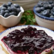A round white plate with blue trim with a serving of blueberry cream cheese dessert on it with two bowls in the background filled with fresh blueberries.