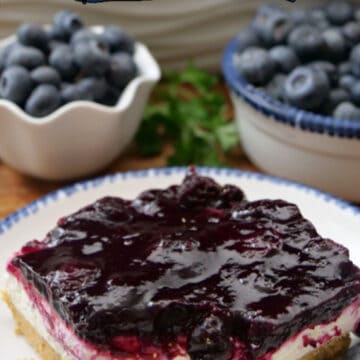 A round white plate with blue trim with a serving of blueberry cream cheese dessert on it with two bowls in the background filled with fresh blueberries.