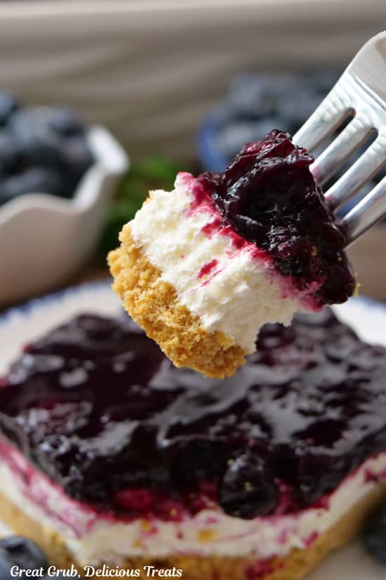 A close up of a bite of blueberry cream cheese dessert on a fork, which has a graham cracker crust, cream cheese mixture and a blueberry topping.