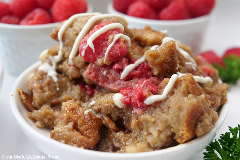 A horizontal photo of a bowl of bread pudding with raspberries and white chocolate drizzle over the top.