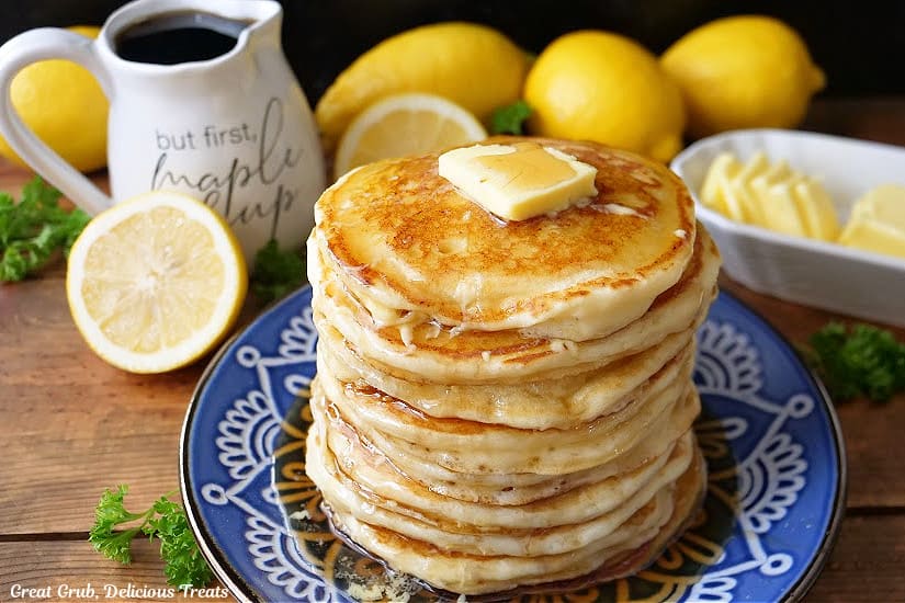 A horizontal photo of a blue plate a stack of eight pancakes on it with fresh lemons, butter, and syrup in the background.