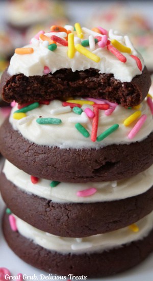 A stack of four brownie cookies with white frosting and jimmie sprinkles and a bite taken out of the top cookie.