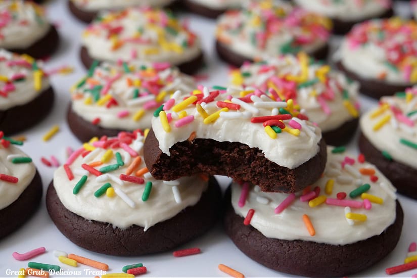 A horizontal photo of a white surface with a couple dozen bite-size brownie cookies with white frosting and jimmie sprinkles on top and a bite taken out of one of the cookies.