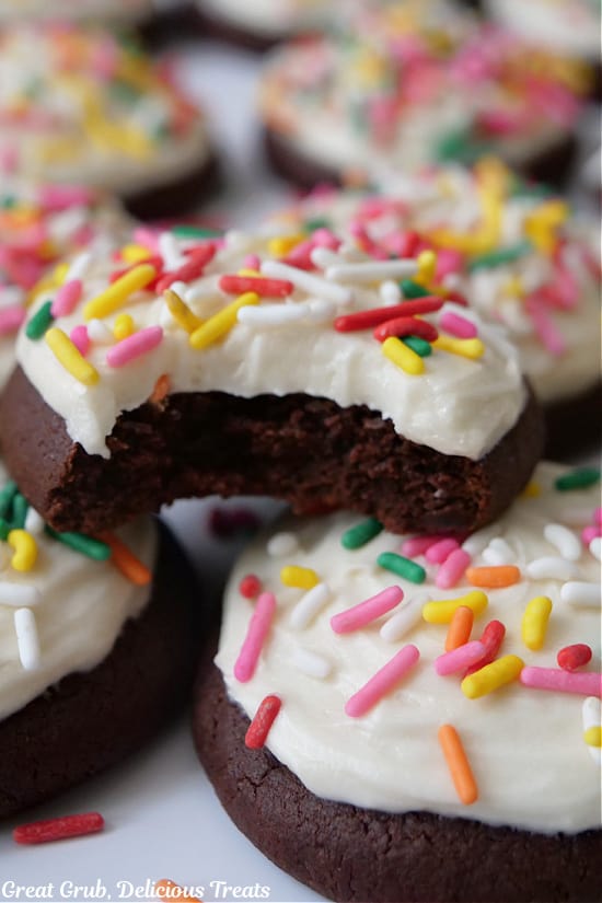 A close up photo of a few brownie cookies with white frosting and candy sprinkles on top and a bite taken out of one of the cookies.