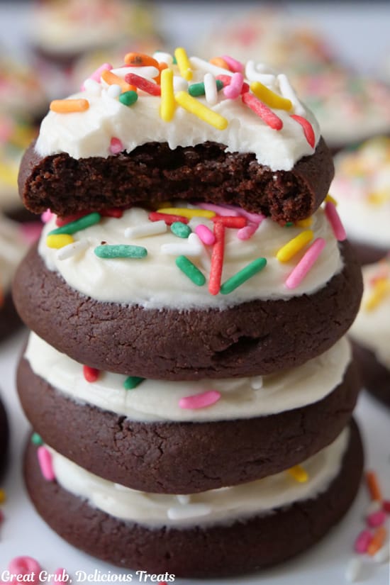 A stack of four brownie cookies that have white frosting and candy sprinkles on top and the top cookie has a bite taken out of it.