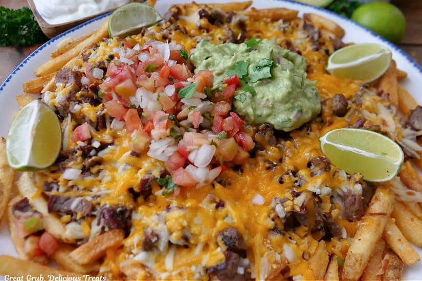 A large plate filled with French fries that are topped with carne asada, melted cheese, Pico de Gallo, guacamole, and has three lime wedges on the plate.