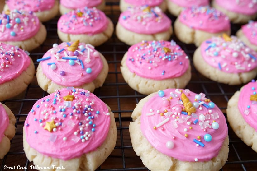 A horizontal photo of 20 bite-size sugar cookies on a wire rack with pink frosting and candy sprinkles on top.